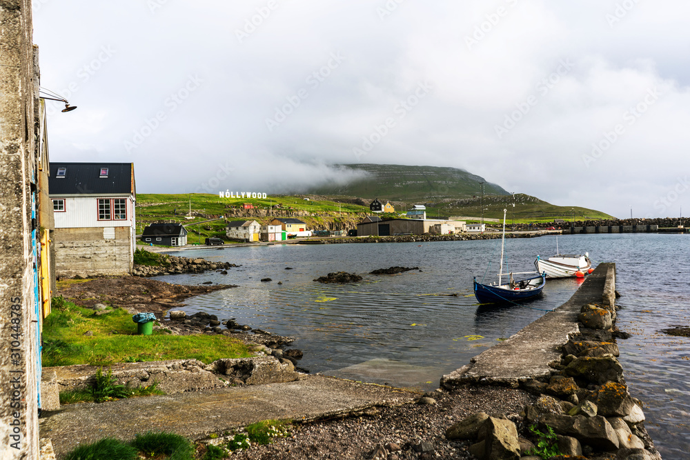 Nolsoy island harbor with boats and small houses, Nollywood sign on a hill. Faroe Islands, Denmark.