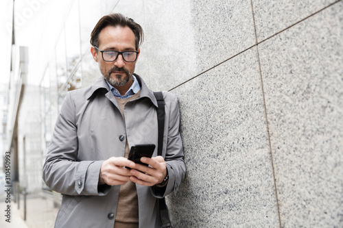Image of serious businessman using cellphone while leaning on wall