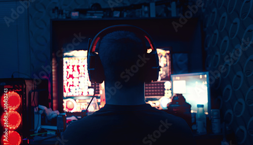 Fototapeta Naklejka Na Ścianę i Meble -  a gamer is sitting in front of a large monitor with headphones, he is an esportsman, a guy of European appearance is shot from the back, a silhouette in headphones against the background of a monitor 