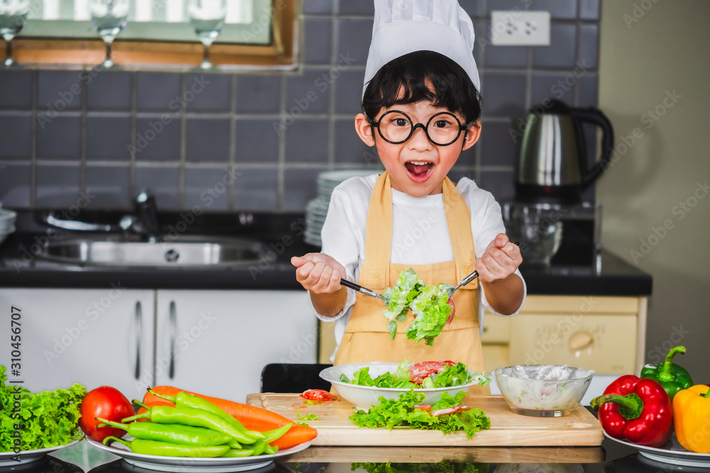 Asian Boy son cooking salad food holdind wooden spoon with vegetable holding tomatoes and carrots, bell peppers on plate for happy family cook food enjoyment lifestyle kitchen in home