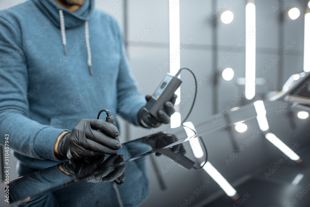 Inspector measuring paint cover thickness of the car body at the vehicle service box, close-up view on hands and professional thickness meter