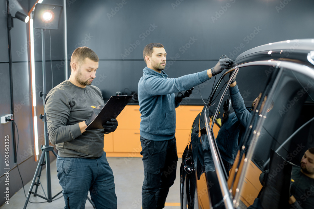 Two car service workers examining vehicle body for scratches and damages while taking a car for professional automotive detailing