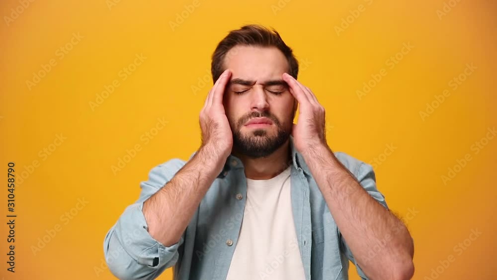 Sad upset bearded young guy 20s in denim shirt white t-shirt isolated over yellow background in studio. People sincere emotions, lifestyle concept. Looks at camera, rubs temples with hands, headache. Sad upset bearded young guy 20s in denim shirt white t-shirt isolated over yellow background in studio. People sincere emotions, lifestyle concept. Looks at camera, rubs temples with hands, headache.