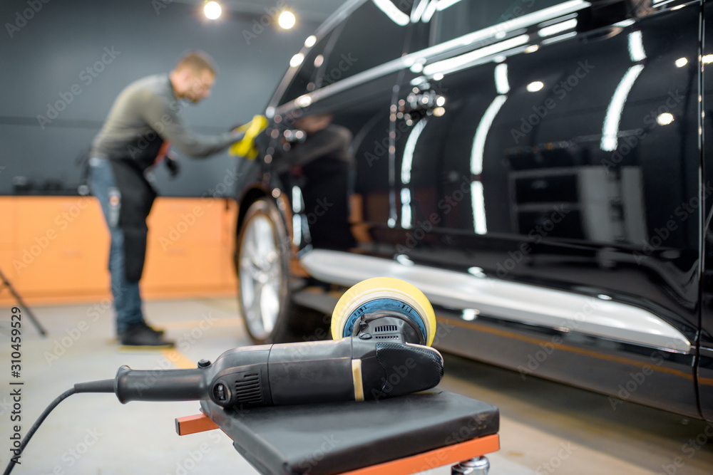 Car service worker wiping vehicle body with microfiber, examining glossy coating after the polishing procedure. Grinder on the foreground