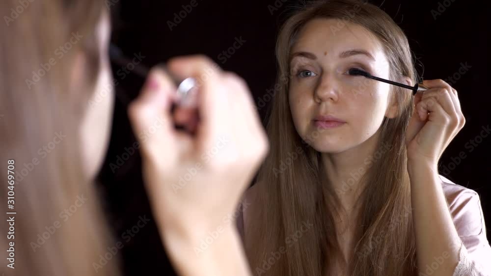 Beautiful Smiling Young Girl Applying Mascara on Eyeleashes and Looking in Mirror at Home. Portrait of Pretty Woman Doing Make Up and Put Cosmetics on Face Beautiful Smiling Young Girl Applying Mascara on Eyeleashes and Looking in Mirror at Home. Portrait of Pretty Woman Doing Make Up and Put Cosmetics on Face