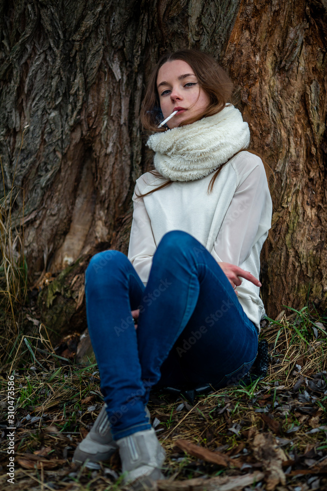 Smoking teenager in real life. Young beautiful white teeage girl in scarf smokes an cigarette near the tree on the street in the autumn. Deadly bad habit. Smoking teenager in real life. Young beautiful white teeage girl in scarf smokes an cigarette near the tree on the street in the autumn. Deadly bad habit.