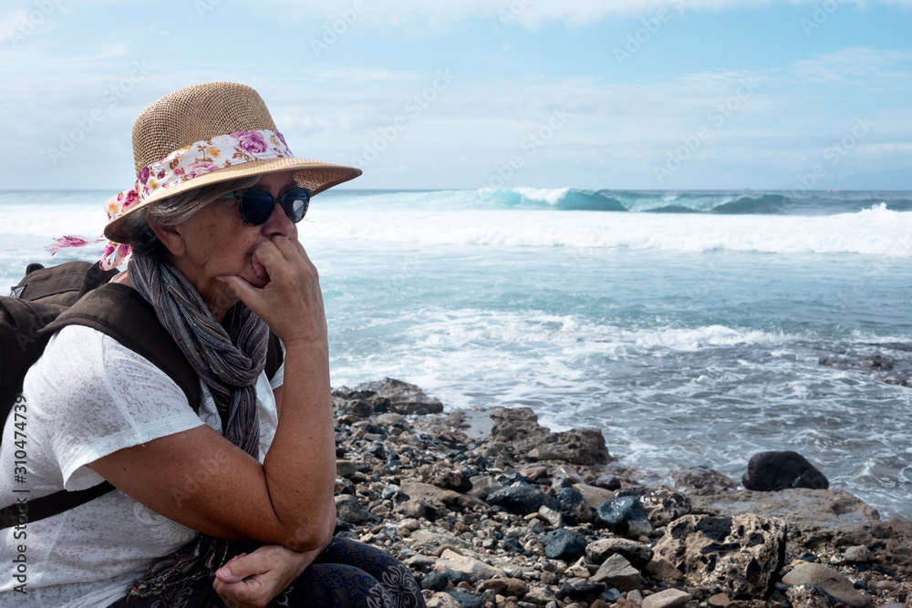 A senior woman with a hat sits thoughtfully on the rocks of the beach in the winter time. Horizon over water. Casual clothing with backpack and hat. Cloudy sky