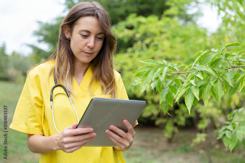 Mujer enfermera joven vestida con uniforme amarillo y estetoscopio colgado del cuello observando con atención la tablet que sostien con ambas manos en entorno de naturaleza Mujer enfermera joven vestida con uniforme amarillo y estetoscopio colgado del cuello observando con atención la tablet que sostien con ambas manos en entorno de naturaleza