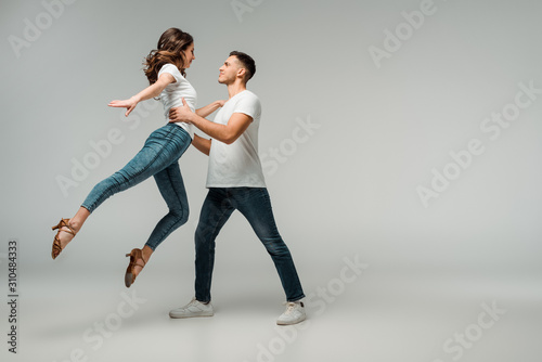side view of smiling dancers in t-shirts and jeans dancing bachata on grey background
