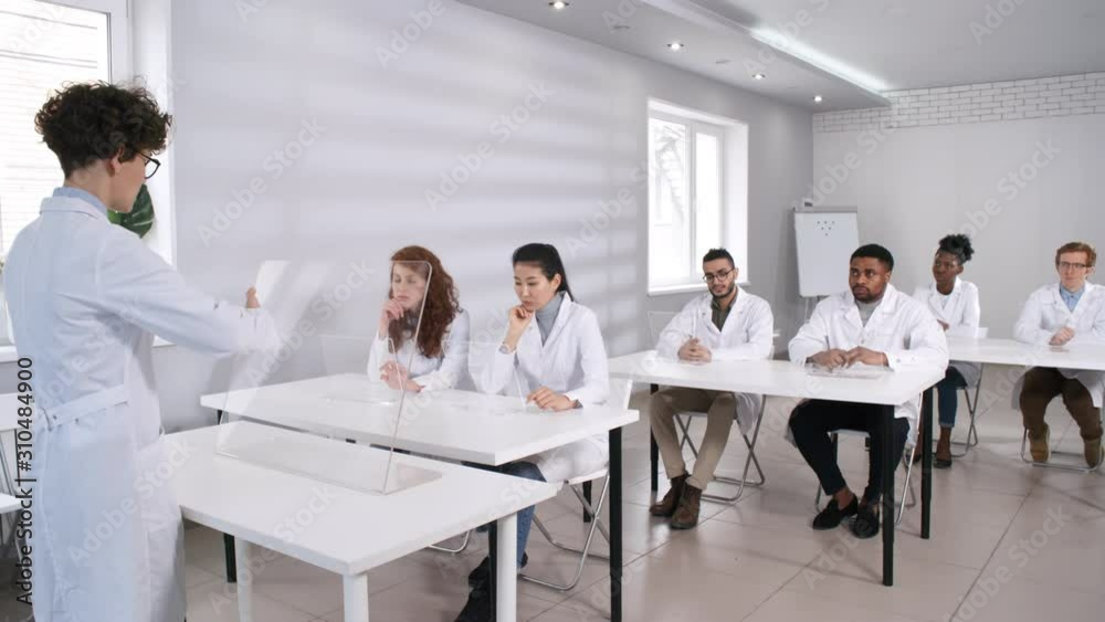 Full shot of female professor in white lab coat and glasses standing in front of multiethnic university students, giving lecture then tapping on interactive transparent AR device. Template for AR, VR. Full shot of female professor in white lab coat and glasses standing in front of multiethnic university students, giving lecture then tapping on interactive transparent AR device. Template for AR, VR.