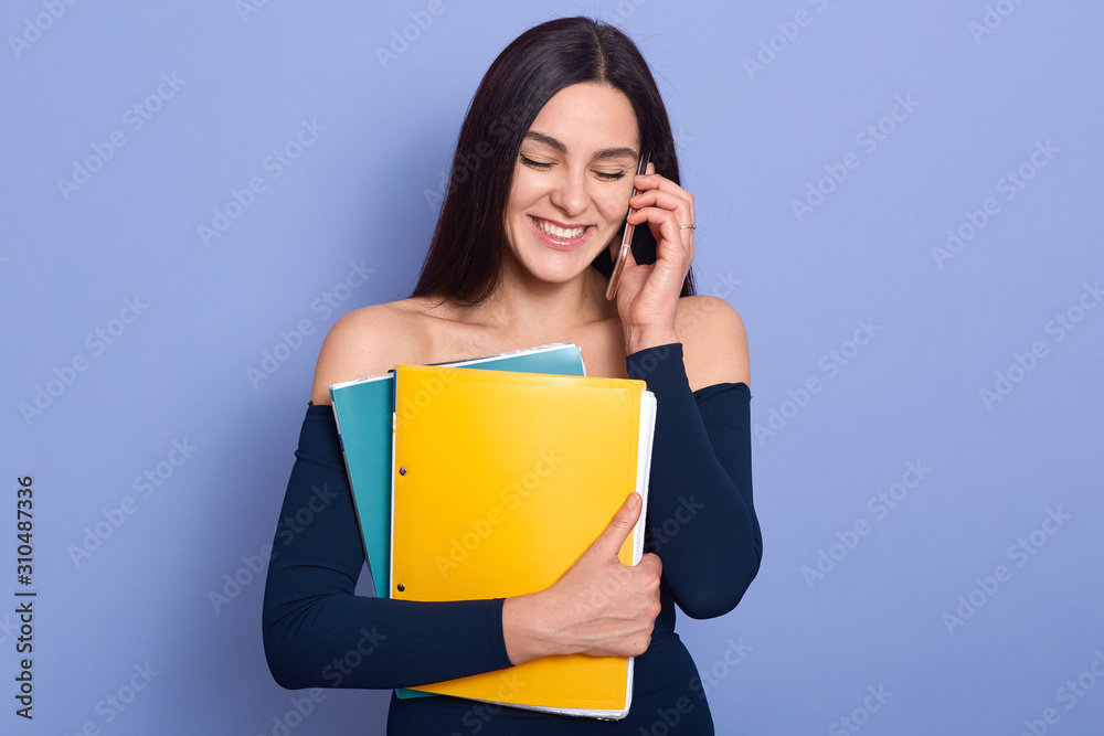 Image of beautiful amazing woman posing over blue background holding paper foldera in hands and talking via mobile phone, looking smiling down, has conversation with boyfriend while having brake. Image of beautiful amazing woman posing over blue background holding paper foldera in hands and talking via mobile phone, looking smiling down, has conversation with boyfriend while having brake.