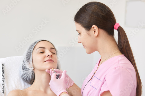 Doctor cosmetologist with ponytail makes rejuvenating facial injections procedure for tightening and smoothing wrinkles on face skin  young woman in beauty salon. Cosmetology and skin care concept.