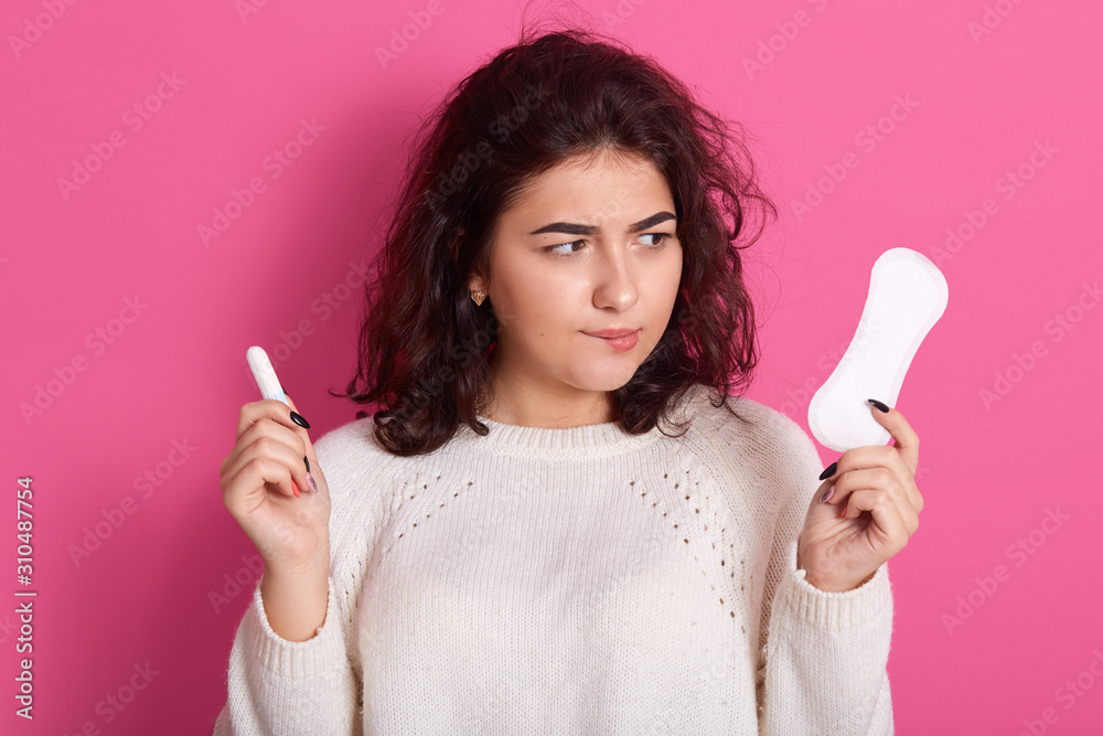 Indoor shot of young brunette woman holding two intimate products, choosing between tampon and pad during menses, thoughtful lady wearing white sweater. Women health care and hygiene concept. Indoor shot of young brunette woman holding two intimate products, choosing between tampon and pad during menses, thoughtful lady wearing white sweater. Women health care and hygiene concept.
