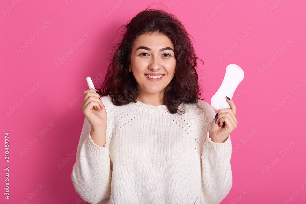 Attractive Caucasian woman holding cotton tampon and sanitary napkin, has happy facial expression, finds best hygiene products for menstrual cycle, lady wearing white sweater. Protective care concept. Attractive Caucasian woman holding cotton tampon and sanitary napkin, has happy facial expression, finds best hygiene products for menstrual cycle, lady wearing white sweater. Protective care concept.