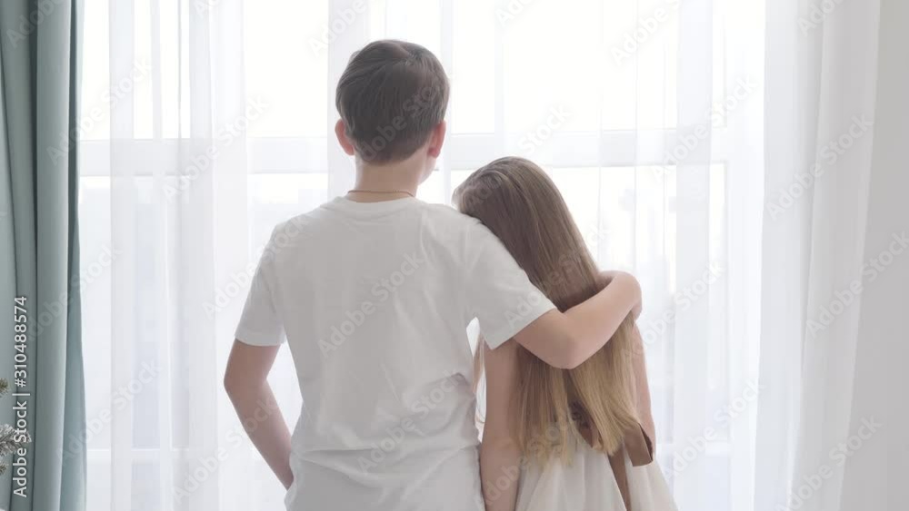 Camera moving away from Caucasian girl and boy standing together in front of the window. Girl's head is on boy's shoulder. First love, happiness. Camera moving away from Caucasian girl and boy standing together in front of the window. Girl's head is on boy's shoulder. First love, happiness.