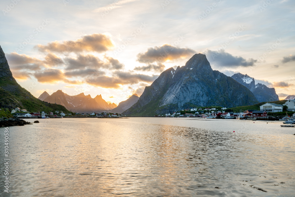 Reine in Lofoten Islands in Norway during sunset. Warm tones and colors fills the cloudy sky during summertime.  Mount olstind in the background. Travelocity and traveling concept.