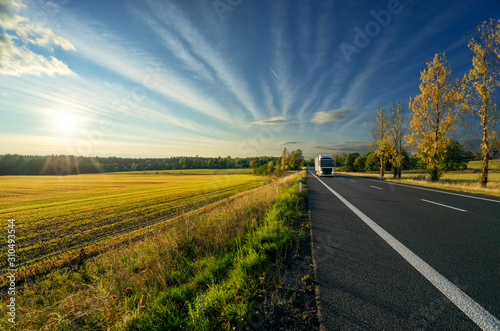 Obraz na płótnie White truck arriving from a distance on an asphalt road in autumn rural landscape at sunset w salonie