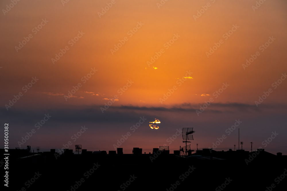 Sun appears from behind the clouds, above the city, roof top view. Summer sunrise or sunset. Orange sky color tones. Old town of Badalona, Spain