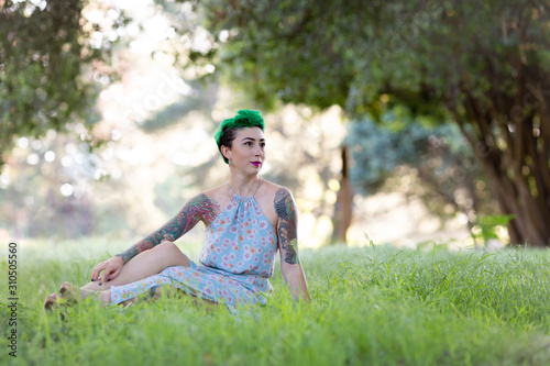 Young Caucasian woman with tattoo and green short hair sitting on green grass meadow in a park, looking to her left © IrinaN