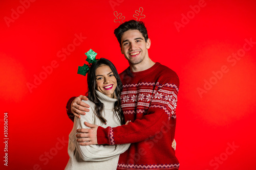 Portrait of young couple in Christmas decorations wreaths on red studio background. Love, holidays, happiness concept.