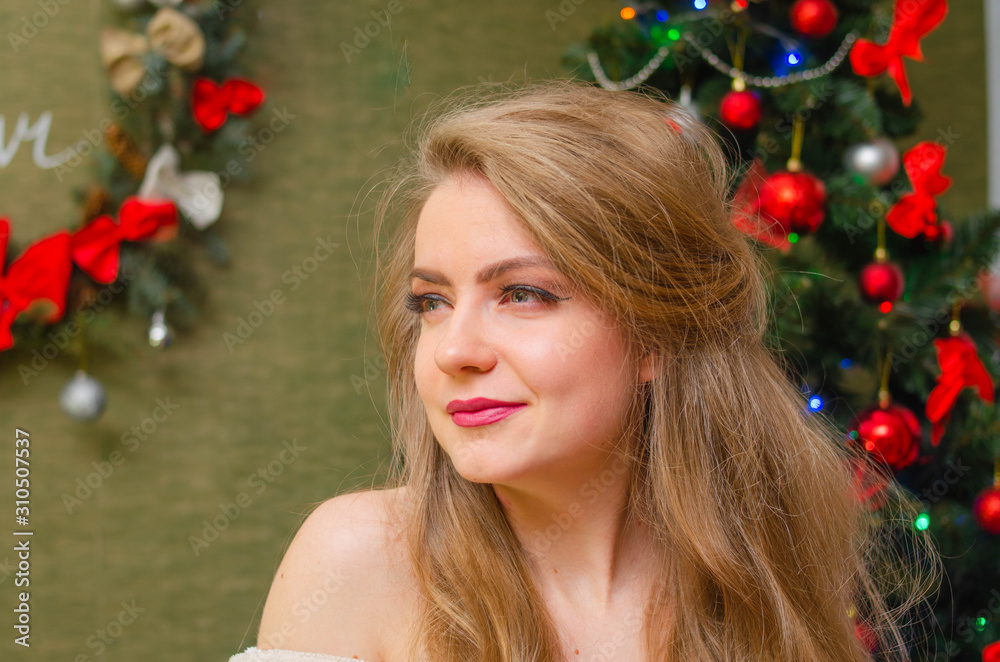 Portrait of a girl with bright red lips, blond long hair against the background of a New Year tree. Young girl in a white warm coat, shoulders are visible. Holidays. Merry Christmas.