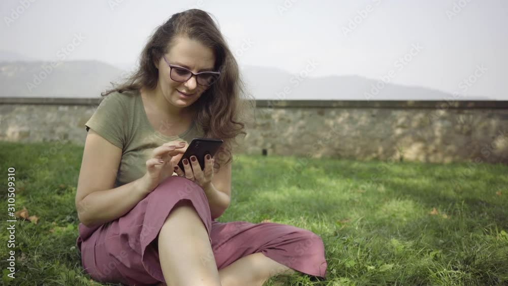 Young woman browsing her phone while seating on the grass in the city park Young woman browsing her phone while seating on the grass in the city park