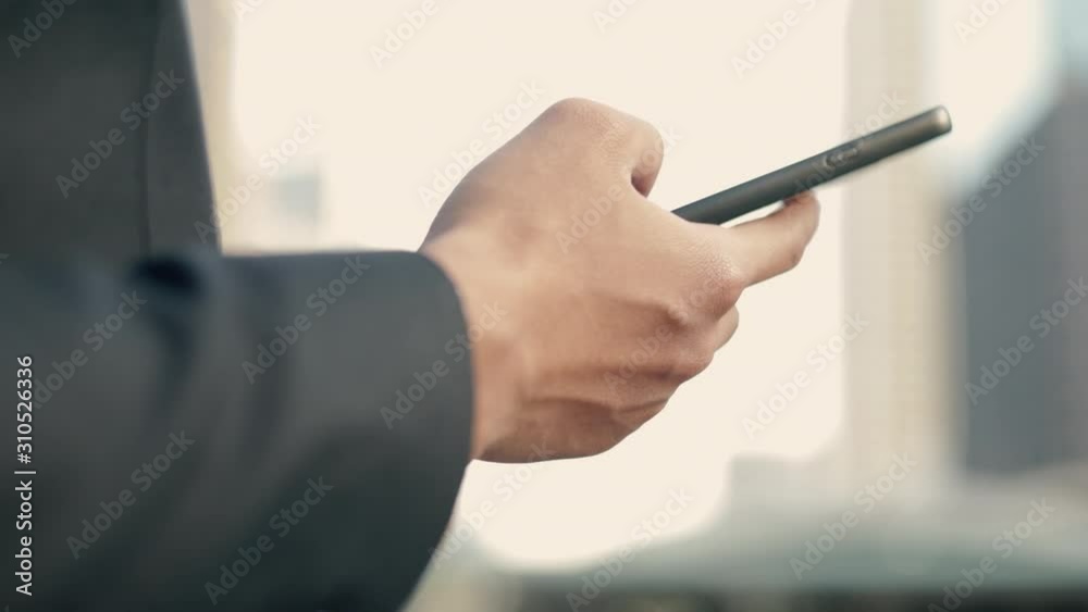 Close up a businessman in a black suit using his smartphone browsing financial, scrolling social media while standing outside on the street near a big office building urban having sunlight sunset. Close up a businessman in a black suit using his smartphone browsing financial, scrolling social media while standing outside on the street near a big office building urban having sunlight sunset.