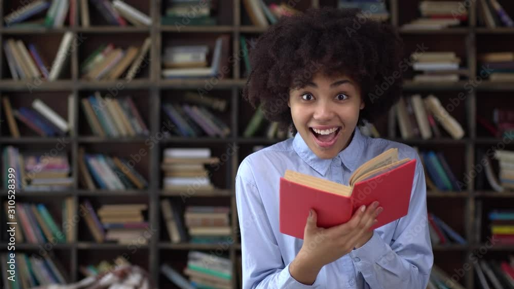 Young african woman in library reading book looking camera cheerful Young african woman in library reading book looking camera cheerful