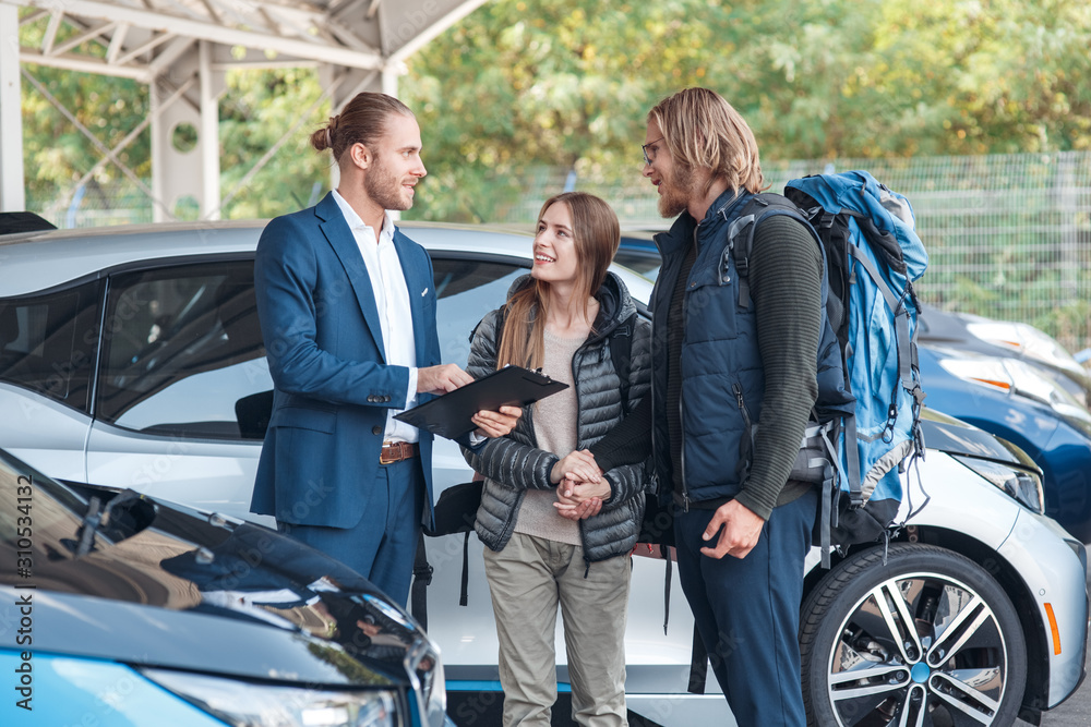 Car Salon. Young couple travelers choosing car for rental with agent holding documents discussing terms and conditions happy Car Salon. Young couple travelers choosing car for rental with agent holding documents discussing terms and conditions happy