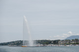 A huge water fountain in the lake with boats in Geneva, Switzerland. Photography of a vertical water stream against a background of a European city in a bay.