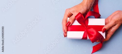 Gift in female hands on a blue background. White box with a red ribbon and red manicure. Banner for Valentine's Day, Christmas or birsday photo