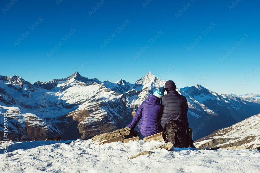 Happy travelers couple man and woman trekking in mountains, enjoy beautiful view. Explorers hiking on snowy hills, travel in Swiss Alps, Switzerland. Romantic hikers sitting outdoors on nature.