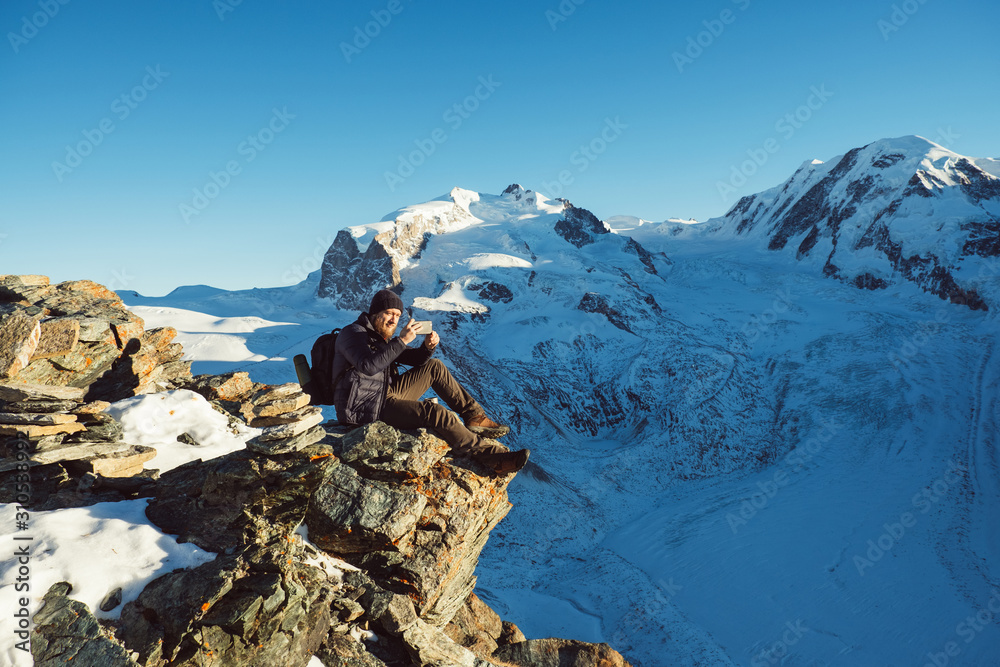 Fototapeta premium Traveler Man with backpack trekking in mountains, enjoy beautiful view and making photo on smartphone. Explorer man hiking, travel in Alps hills, Switzerland. Hiker sitting on rock cliff outdoors.