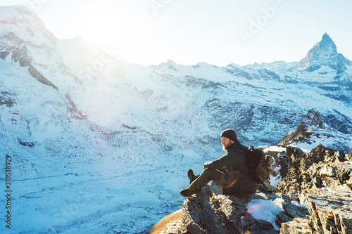 Traveler Man with backpack trekking in mountains, enjoy beautiful Matterhorn view. Explorer man hiking on snowy hills, travel in Alps, Switzerland. Hiker sitting on rock cliff outdoors on nature.