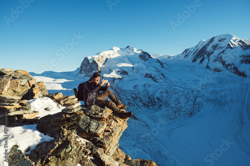 Traveler Man with backpack trekking in mountains  enjoy beautiful view and making photo on smartphone. Explorer man hiking  travel in Alps hills  Switzerland. Hiker sitting on rock cliff outdoors.