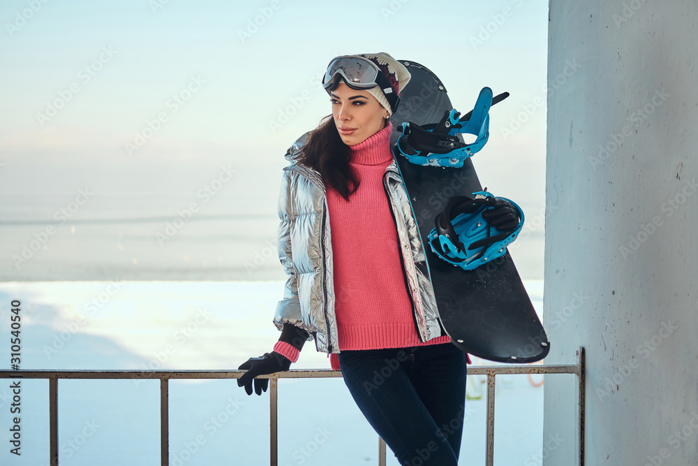 Obraz premium Young active woman with snowboard is posing for photographer at bright winter day.