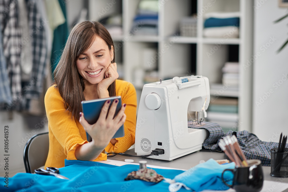 Smiling positive caucasian brunette sitting in her fashion studio and using tablet.