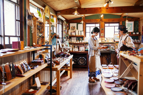 Japanese woman wearing apron standing in a leather shop, holding tan leather clutch bag, talking to customer. photo