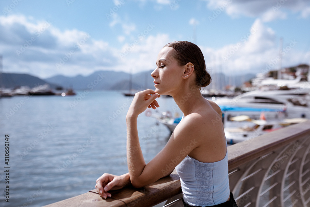 Young tourist woman on a bridge. The girl in a vest is in a thoughtful mood. Woman looks dreamy on the port with ships. Seascape with mountains. Summer vacation in Marmaris, Turkey. Copy space.