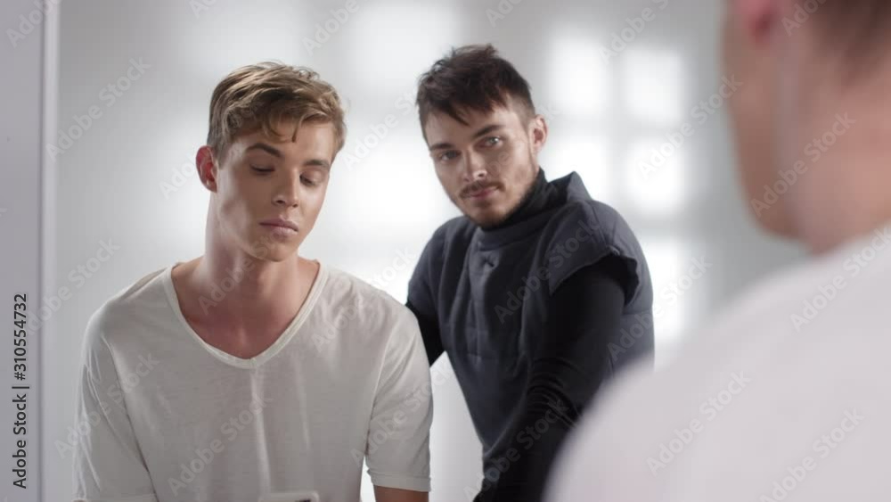 Back view of young man with blonde hair sitting in front of mirror in makeup studio while male visagiste coloring his brows. Then model making selfie on smartphone camera Back view of young man with blonde hair sitting in front of mirror in makeup studio while male visagiste coloring his brows. Then model making selfie on smartphone camera