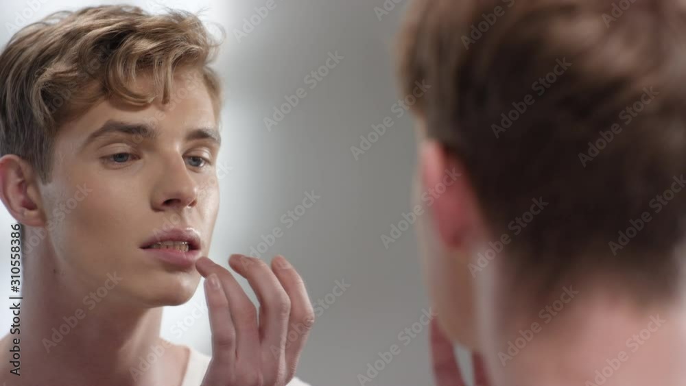 Back view of young blond-haired Caucasian man standing in front of mirror and applying lip balm with his finger Back view of young blond-haired Caucasian man standing in front of mirror and applying lip balm with his finger