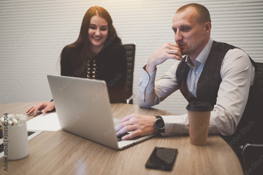 Two business partners in a meeting room (a caucasian man entrepreneur with a smartphone and a paper cup, and a businesswoman) having a video conference with their colleagues via laptop on the table Two business partners in a meeting room (a caucasian man entrepreneur with a smartphone and a paper cup, and a businesswoman) having a video conference with their colleagues via laptop on the table