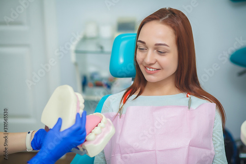 Young dentist holding a plastic model of teeth and explaining the procedure of new dental implants to her patient sitting in dental chair at stomatology