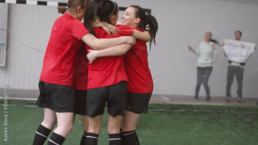 Group of young joyous female players jumping, huddling and giving high five to each other while celebrating soccer goal during match on indoor sports field