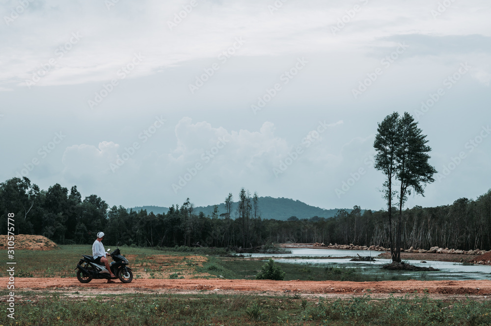 Fototapeta premium Man wearing a helmet is having a rest next to the lake with the high tree in the middle after riding a motorbike on the Phu Quoc island in Vietnam. Cloudy grey sky before the rain.