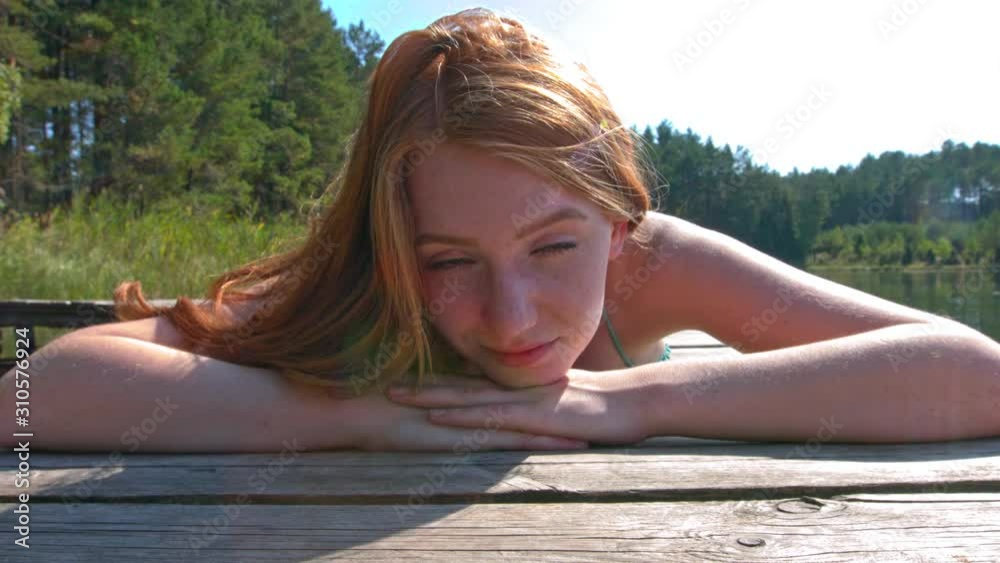 A close-up of a woman's face resting on her hands, as she lays down on a wooden jetty looking into the camera, soaking up the midday sun