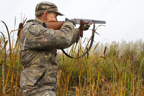 Photo hunter aiming a shotgun among cattail