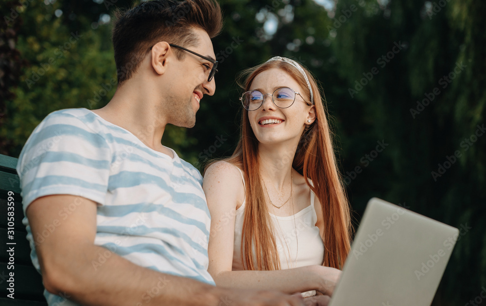 Portrait of a charming young couple looking to each other laughing while man is holding a laptop on his legs freelancing outdoor in park on a bench. Portrait of a charming young couple looking to each other laughing while man is holding a laptop on his legs freelancing outdoor in park on a bench.