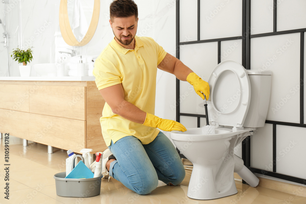 Young man feeling disgust while cleaning toilet bowl in bathroom - obrazy, fototapety, plakaty 