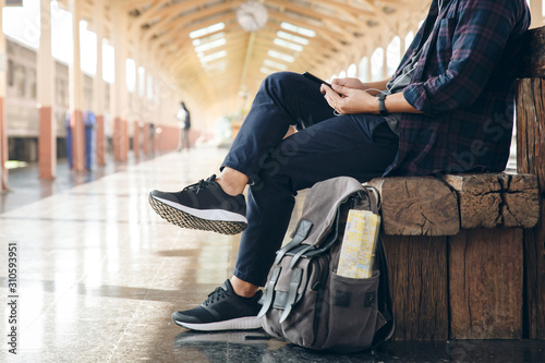 Young man traveler sitting with using mobile phone choose where to travel and bag waiting for train at train station.backpacker at the train station and looking on mobile phone for plan to travel.